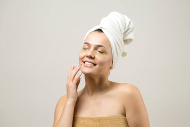 Kafasında beyaz havluyla gezen genç bir kız gözlerinin altına kolajen jel bantlar takıyor. Göz teması altında maske.