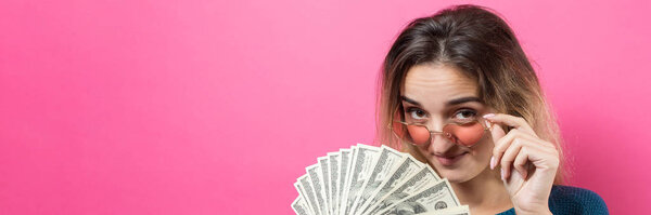 Closeup of young beautiful woman in a blue sweater in glasses with american dollars money in hand over pink background. Expresses emotions and gestures of joy.