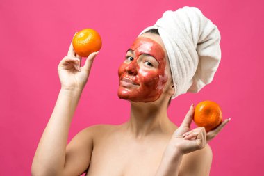 Kafasında kırmızı besleyici maske olan beyaz havluyla güzel bir kadın portresi. Cilt temizleme organik kozmetik rahatlama konsepti. Sırtı turuncu bir mandalina tutan bir kız..