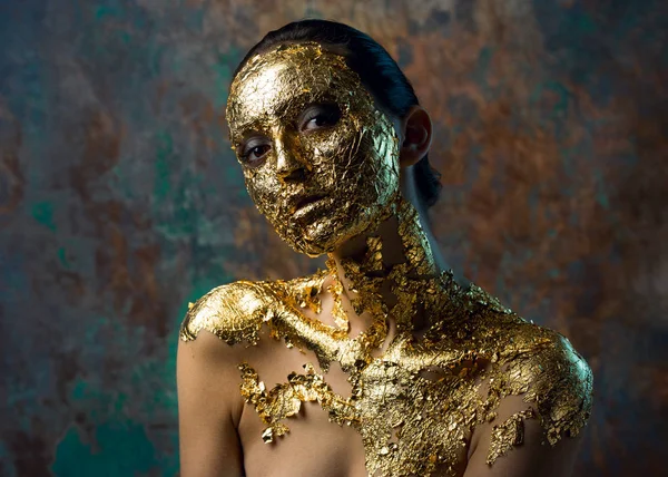 Girl with a mask on her face made of gold leaf. Gloomy studio portrait of a brunette on an abstract background.