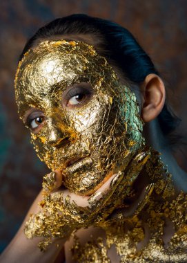 Yüzünde altın yapraktan yapılmış bir maske olan bir kız. Soyut bir arka planda esmer bir kadının kasvetli stüdyo portresi.