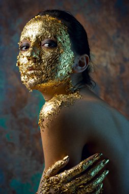Yüzünde altın yapraktan yapılmış bir maske olan bir kız. Soyut bir arka planda esmer bir kadının kasvetli stüdyo portresi.