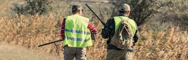 A mans with a gun in his hands and an camouflage vest on a pheasant hunt in a wooded area in cloudy weather. Hunters in search of game.