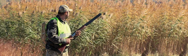 A man with a gun in his hands and an green vest on a pheasant hunt in a wooded area in cloudy weather. Hunter with dogs in search of game.