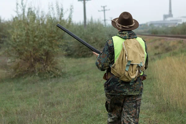 A man with a gun in his hands and an orange vest on a pheasant hunt in a wooded area in cloudy weather. Hunter with dogs in search of game.