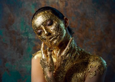 Yüzünde altın yapraktan yapılmış bir maske olan bir kız. Soyut bir arka planda esmer bir kadının kasvetli stüdyo portresi.