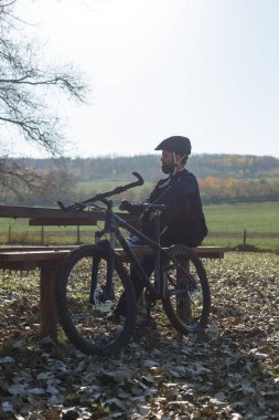 Pantolon ve yün ceketli bir bisikletçi modern karbon donanımlı bir motorsikletle off-road 'a biniyor. Adam sonbahar parkında bir bankta dinleniyor..
