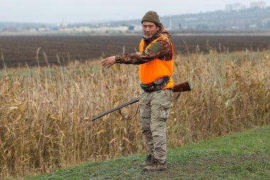 Elinde silah ve turuncu yelek olan bir adam bulutlu bir havada ormanlık alanda sülün avında. Avcı köpeklerle av peşinde.