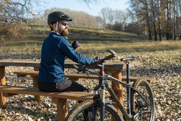 Cyclist in pants and fleece jacket on a modern carbon hardtail bike with an air suspension fork rides off-road. The guy is resting on a bench in the autumn park.