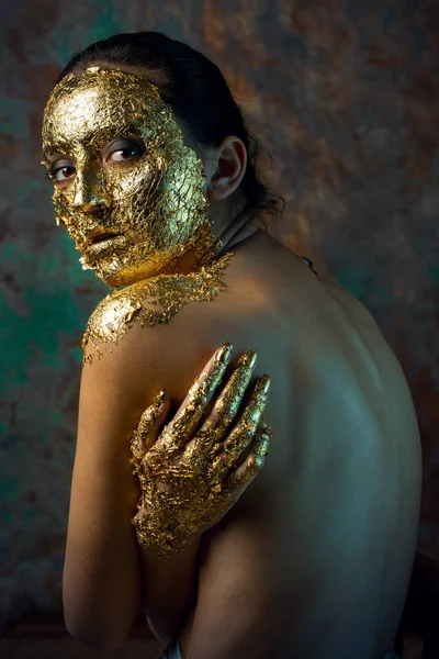 Girl with a mask on her face made of gold leaf. Gloomy studio portrait of a brunette on an abstract background.