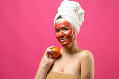 Kafasında kırmızı besleyici maske olan beyaz havluyla güzel bir kadın portresi. Cilt temizleme organik kozmetik rahatlama konsepti. Sırtı turuncu bir mandalina tutan bir kız..