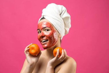 Kafasında kırmızı besleyici maske olan beyaz havluyla güzel bir kadın portresi. Cilt temizleme organik kozmetik rahatlama konsepti. Sırtı turuncu bir mandalina tutan bir kız..