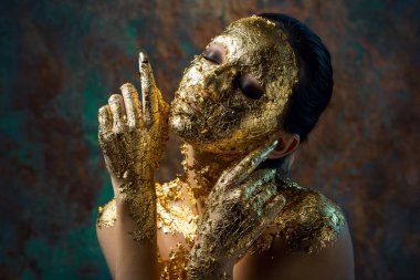 Yüzünde altın yapraktan yapılmış bir maske olan bir kız. Soyut bir arka planda esmer bir kadının kasvetli stüdyo portresi.