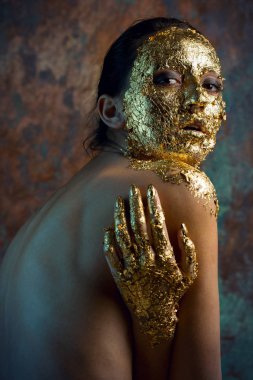 Yüzünde altın yapraktan yapılmış bir maske olan bir kız. Soyut bir arka planda esmer bir kadının kasvetli stüdyo portresi.