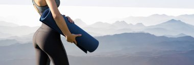 Spor giyimli genç ve zayıf bir atletik kız, dağların arka planında bir takım egzersizler yapıyor. Yoga minderiyle duruyor..