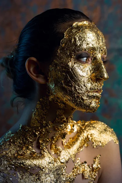 Girl with a mask on her face made of gold leaf. Gloomy studio portrait of a brunette on an abstract background.