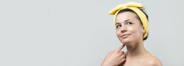 Kafasında beyaz havluyla gezen genç bir kız gözlerinin altına kolajen jel bantlar takıyor. Göz teması altında maske.