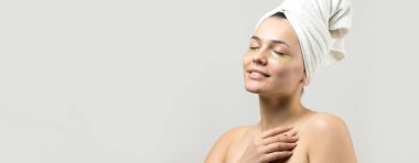 Kafasında beyaz havluyla gezen genç bir kız gözlerinin altına kolajen jel bantlar takıyor. Göz teması altında maske.