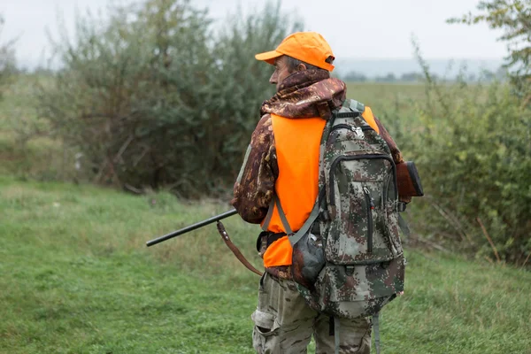 A man with a gun in his hands and an orange vest on a pheasant hunt in a wooded area in cloudy weather. Hunter with dogs in search of game.