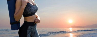 Spor giyimli genç ve zayıf bir atletik kız deniz kenarında günbatımının arka planında bir dizi egzersiz yapıyor. Yoga minderiyle duruyor..