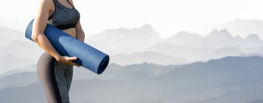 Spor giyimli genç ve zayıf bir atletik kız, dağların arka planında bir takım egzersizler yapıyor. Yoga minderiyle duruyor..