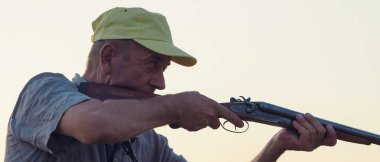 Elinde silah ve yeşil yelek olan bir adam bulutlu bir havada ormanlık alanda sülün avında. Avcı köpeklerle av peşinde.