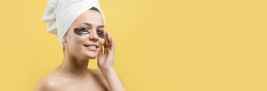 Kafasında beyaz havluyla gezen genç bir kız gözlerinin altına kolajen jel bantlar takıyor. Göz teması altında maske.