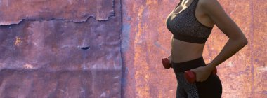 Spor giyimli genç, zayıf, atletik bir kız paslı arka planlar üzerinde bir takım egzersizler yapıyor. Dambıllarla egzersiz yapar. Fitness ve sağlıklı yaşam tarzı.