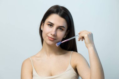 Açık renk bluzlu, beyaz arka planda yüz makyajı yapan genç bir kızın yakın çekimi. Güzel bir kadın yüzüne bir makyaj fırçası tutar ve gülümser..