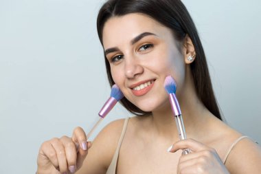 Açık renk bluzlu, beyaz arka planda yüz makyajı yapan genç bir kızın yakın çekimi. Güzel bir kadın yüzüne bir makyaj fırçası tutar ve gülümser..