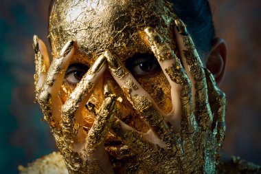 Yüzünde altın yapraktan yapılmış bir maske olan bir kız. Soyut bir arka planda esmer bir kadının kasvetli stüdyo portresi.