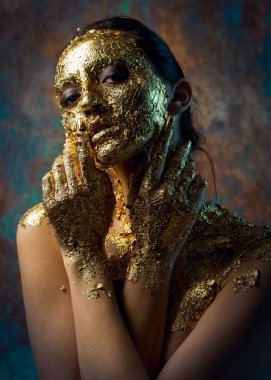 Yüzünde altın yapraktan yapılmış bir maske olan bir kız. Soyut bir arka planda esmer bir kadının kasvetli stüdyo portresi.