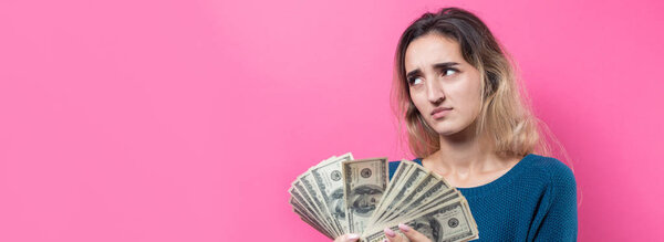 Closeup of young beautiful woman in a blue sweater in glasses with american dollars money in hand over pink background. Expresses emotions and gestures of joy.