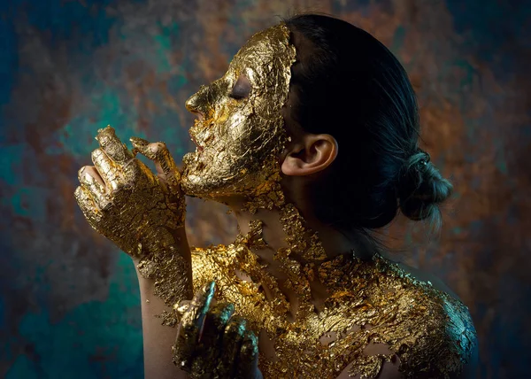 Girl with a mask on her face made of gold leaf. Gloomy studio portrait of a brunette on an abstract background.