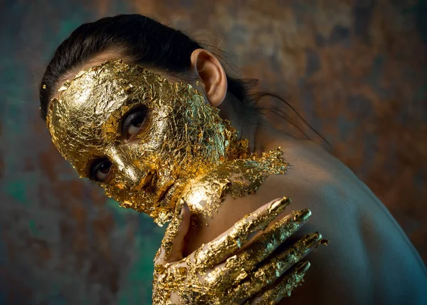 Girl with a mask on her face made of gold leaf. Gloomy studio portrait of a brunette on an abstract background.