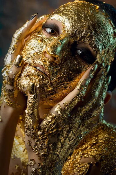 Girl with a mask on her face made of gold leaf. Gloomy studio portrait of a brunette on an abstract background.