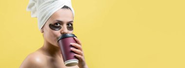 Kafasında beyaz havluyla gezen genç bir kız gözlerinin altına kolajen jel bantlar takıyor. Göz tedavisiyle maskelenmiş. Kız elinde bir bardak kahve tutuyor..