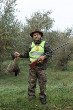 Elinde silah ve yeşil yelek olan bir adam bulutlu bir havada ormanlık alanda sülün avında. Avcı köpeklerle av peşinde.