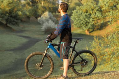 Taze yeşil yaz ormanının arka planına karşı bir uçurumun üzerinde duran bir hava süspansiyon çatal ile modern bir karbon hardtail bisiklet üzerinde şort ve mayo Bisikletçi