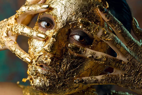 Girl with a mask on her face made of gold leaf. Gloomy studio portrait of a brunette on an abstract background.