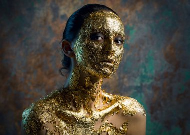 Yüzünde altın yapraktan yapılmış bir maske olan bir kız. Soyut bir arka planda esmer bir kadının kasvetli stüdyo portresi.