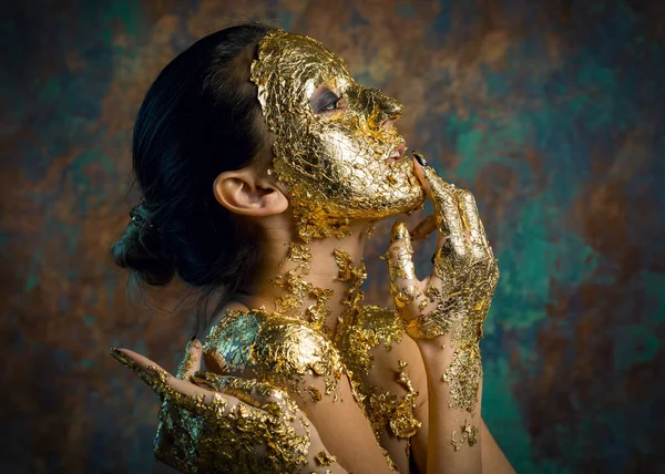 Girl with a mask on her face made of gold leaf. Gloomy studio portrait of a brunette on an abstract background.