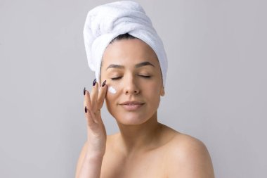 Kafasında beyaz havlu olan bir kadının güzel portresi yüzüne krem sürüyor. Cilt temizleme eko organik kozmetik rahatlama konsepti.