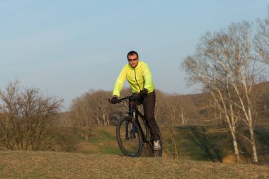 Pantolon ve yeşil ceketli bir bisikletçi. Modern karbon donanımlı, çatallı bir motosiklet. Tepedeki adam bisiklete biniyor..