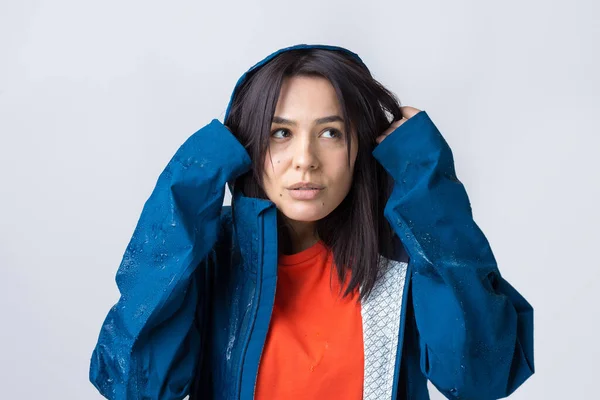 Portrait of a smiling girl dressed in blue raincoat in drops posing with hood on grey background in a studio.