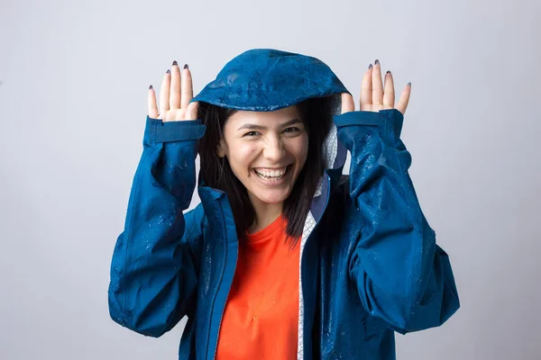 Portrait of a smiling girl dressed in blue raincoat in drops posing with hood on grey background in a studio.