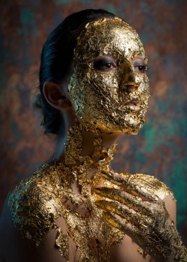 Yüzünde altın yapraktan yapılmış bir maske olan bir kız. Soyut bir arka planda esmer bir kadının kasvetli stüdyo portresi.