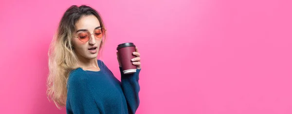 ピンクの背景にコーヒーを飲みながら青いセーターを着たグラマーな女性 — ストック写真