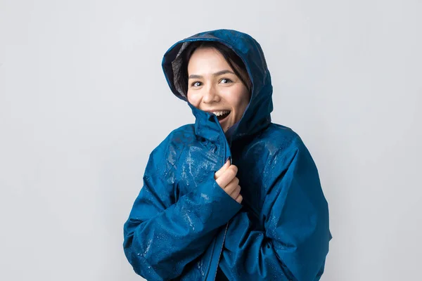 Portrait of a smiling girl dressed in blue raincoat in drops posing with hood on grey background in a studio.