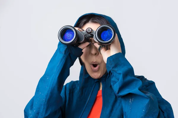 A tourist girl in a blue raincoat holds binoculars in her hands and looks into the distance, spies.